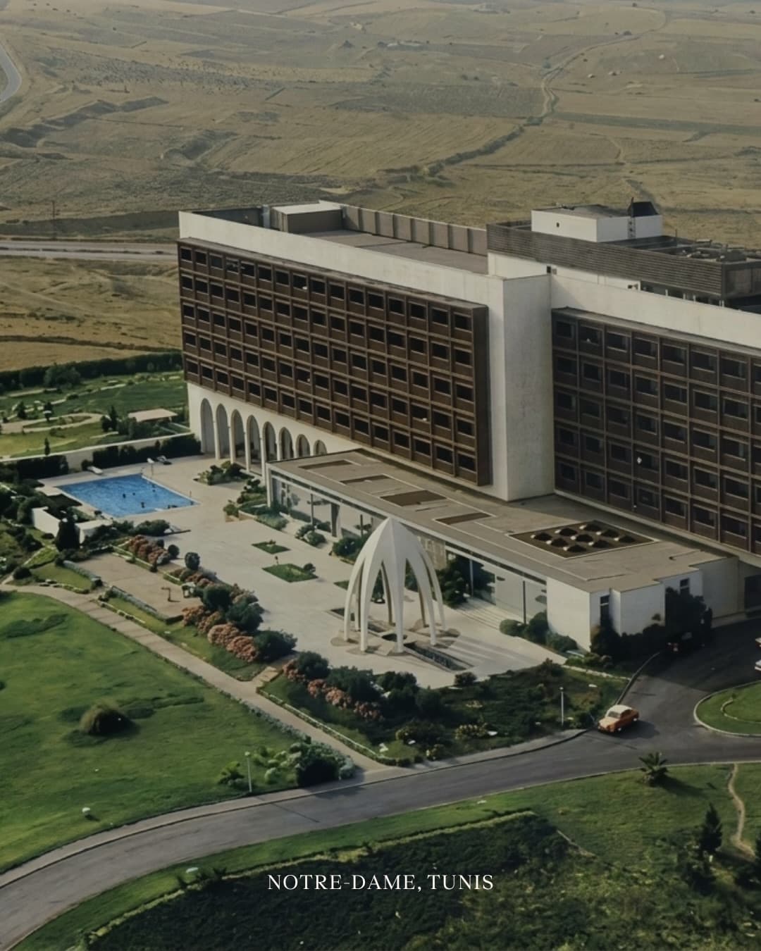 Vue aérienne d’un hôtel à la façade quadrillée, avec piscine, jardins et une entrée surmontée d’arches sculptées ; mention « Notre-Dame, Tunis ».